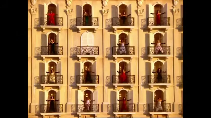 A large group of women in colorful dresses appear on balconies of a grand building, then disappear, in a dramatic and synchronized fashion.