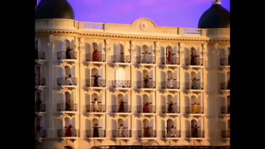 A wide shot of a grand hotel facade with many women in colorful dresses appearing on balconies, against a purple sky.