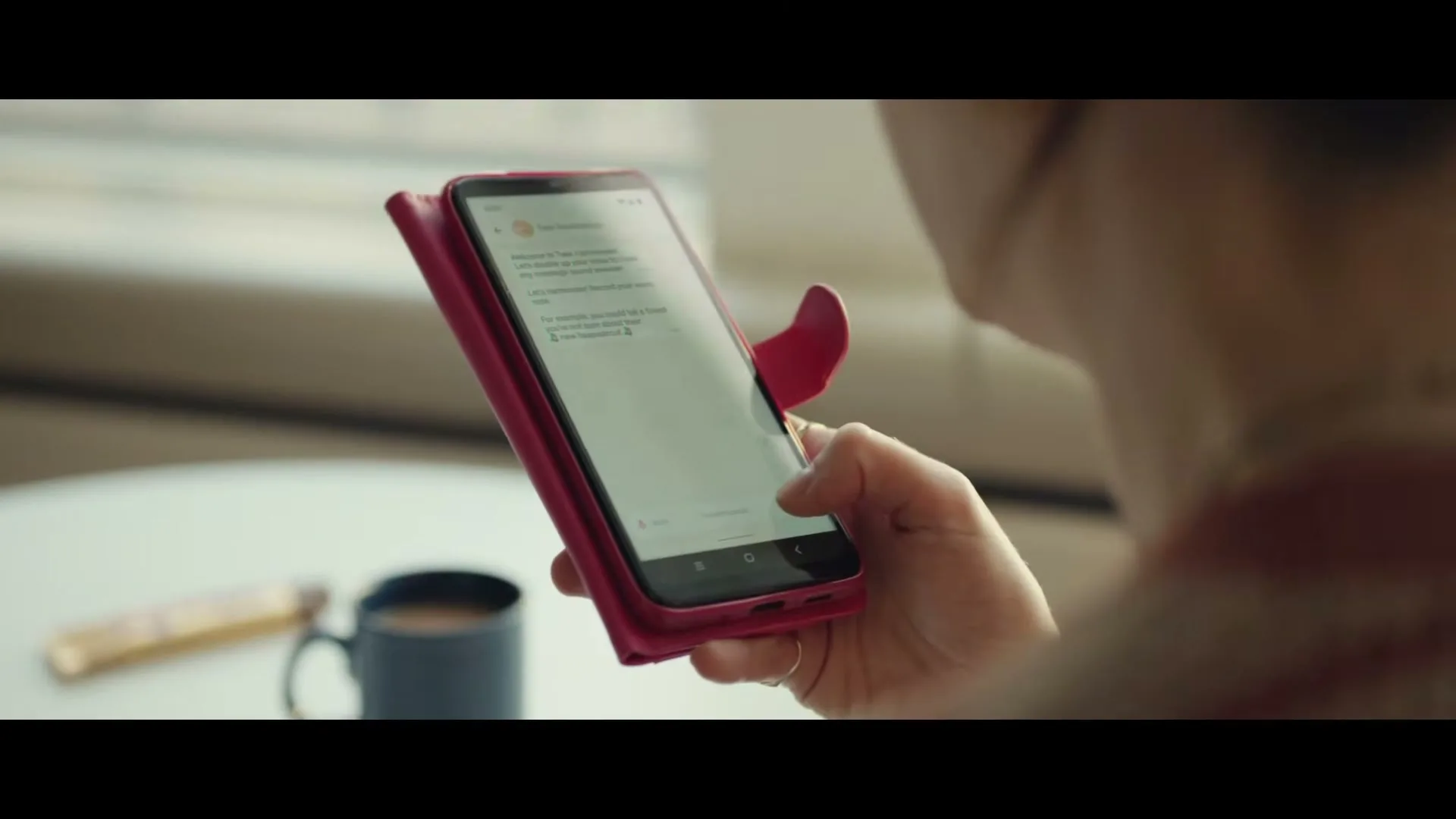 A woman holds a smartphone with a pink case, interacting with an app, while a Twix bar and a mug are visible on a table in the background.