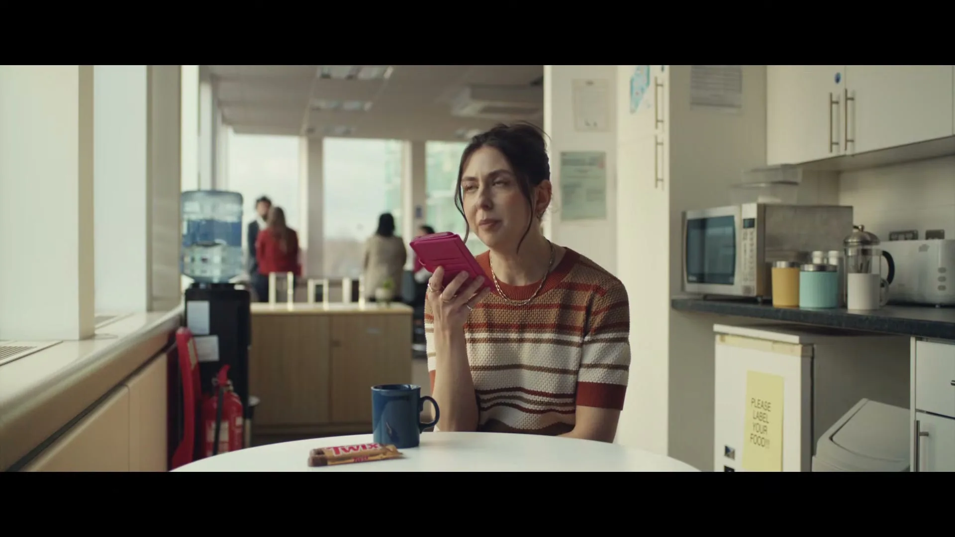 A woman in a striped shirt sits at a table in an office kitchen, holding a pink phone to her ear and looking concerned.