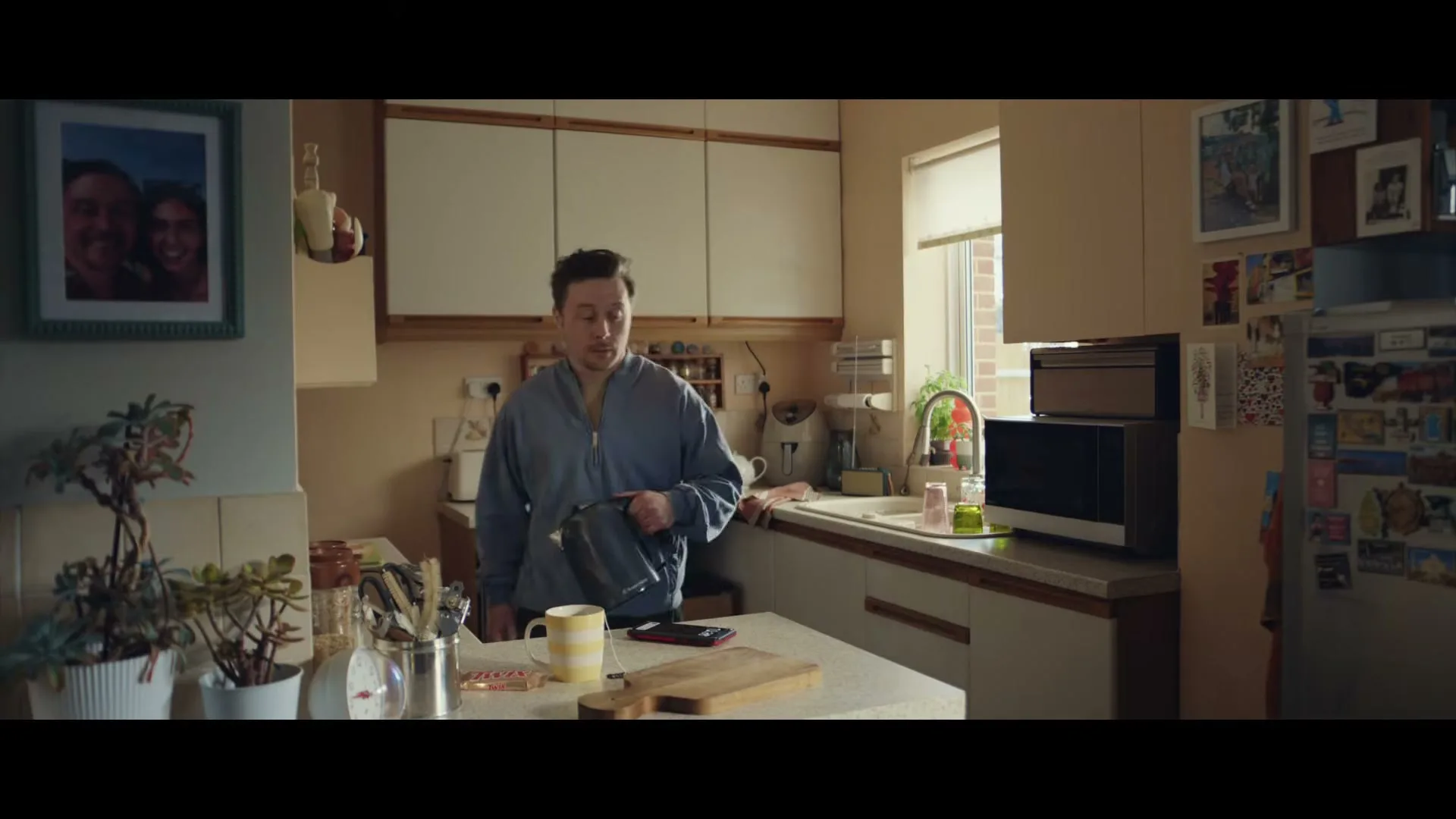 A man in a blue sweatshirt walks into a cluttered kitchen, holding a lunchbox, and places it on the counter.