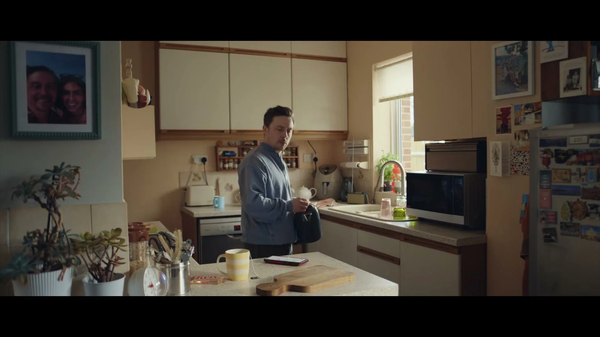 A man in a blue sweater stands in a kitchen, looking at something in his hand, with a mug and phone on the counter.