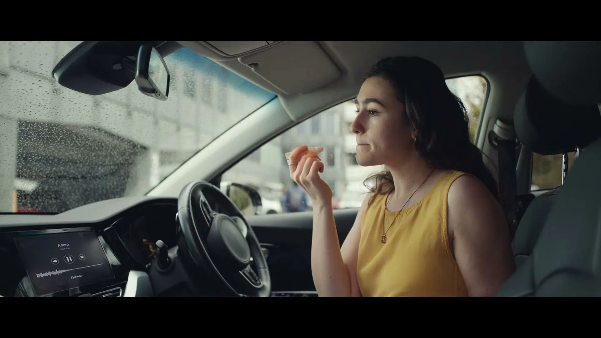 A young woman in a yellow top applies lip balm while sitting in the driver's seat of a car during the day.