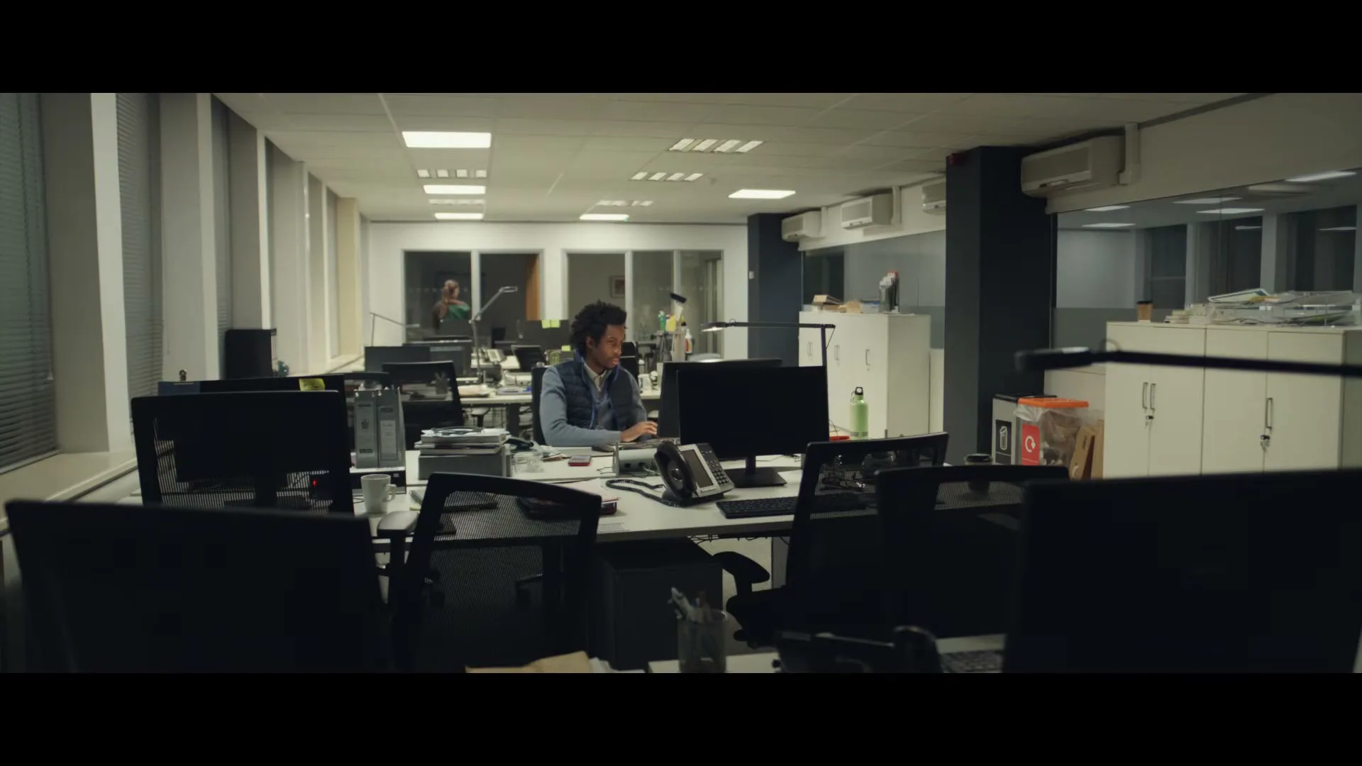 A man with curly hair in a blue vest sits at a desk in a dimly lit office, typing on a computer.