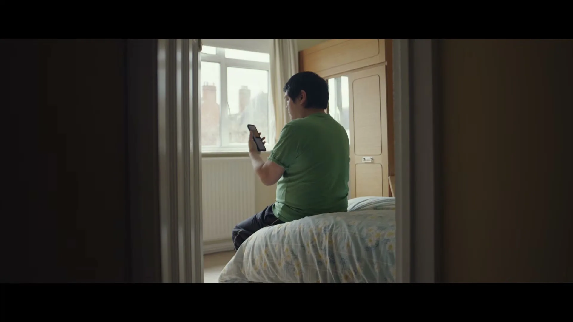 A man in a green t-shirt sits on a bed, looking at his phone, with a window and wardrobe in the background, seen through a doorway.