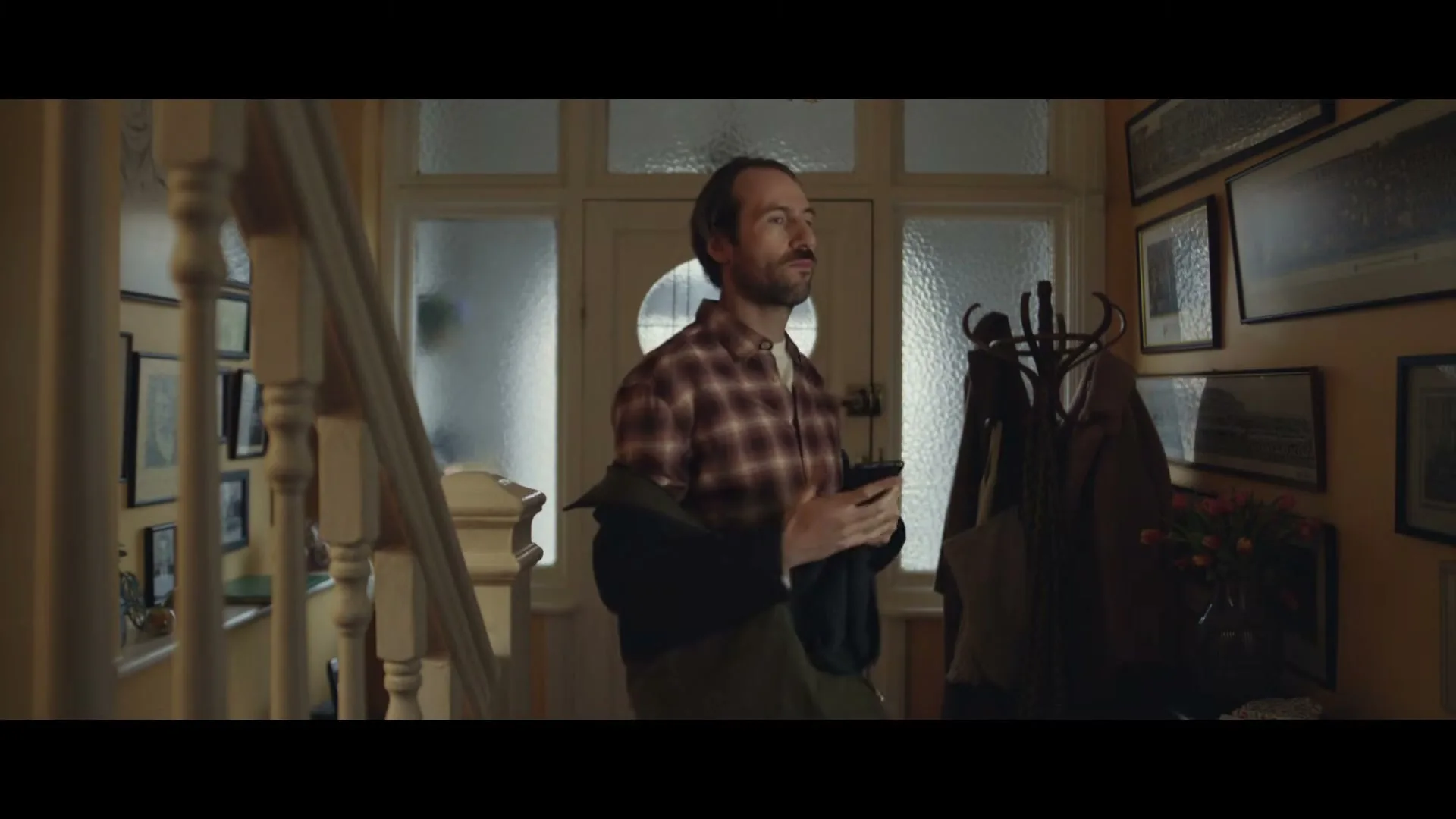 A man in his 30s, wearing a plaid shirt, stands in a home entryway, holding a phone and looking concerned as he takes off his coat.
