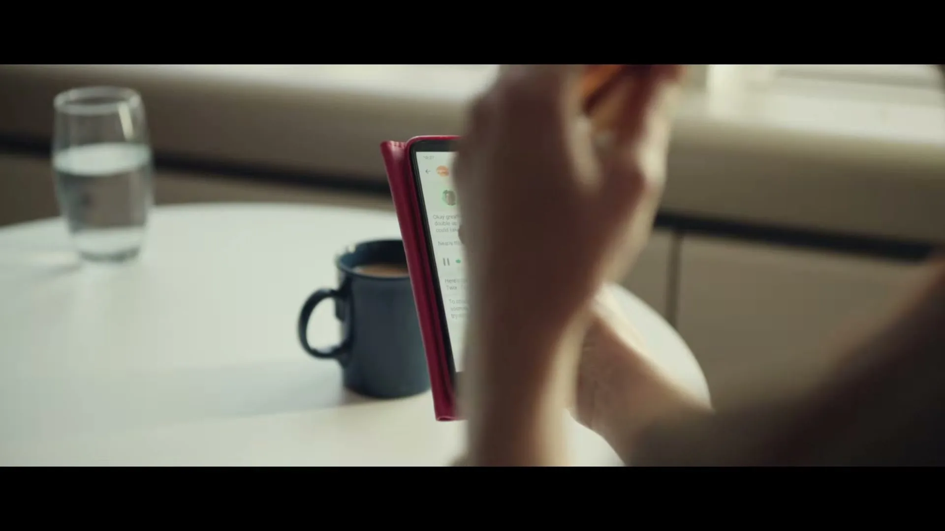 A person's hands hold a Twix bar and a smartphone displaying a messaging app, with a coffee mug and water glass on a table in the background.