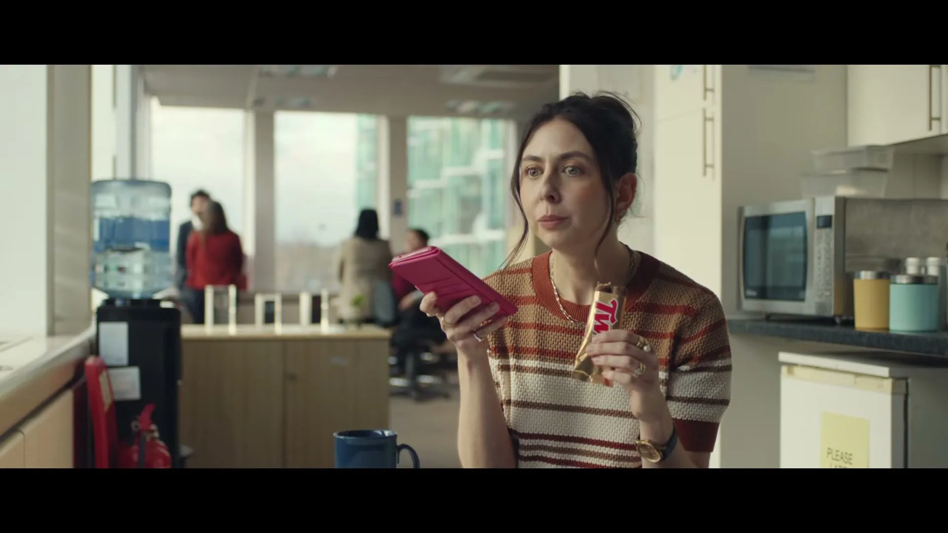 A woman in her 30s, wearing a striped sweater, holds a pink phone and eats a Twix bar in an office kitchen.