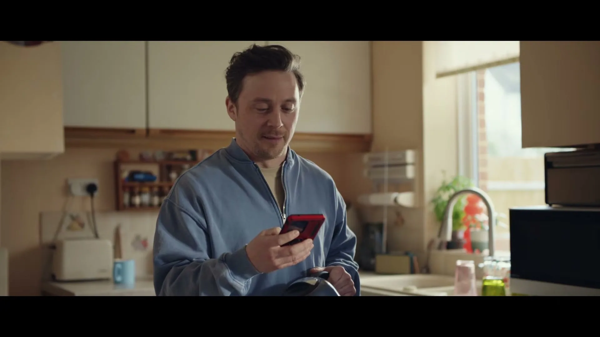 A man in his 30s, wearing a blue jacket, stands in a kitchen, looking at his red phone and smiling, with text on screen.