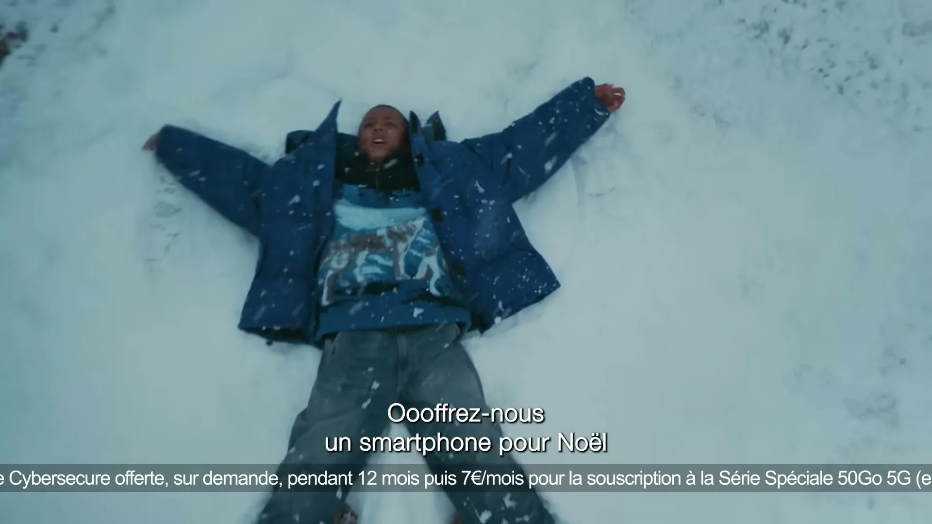 A child in a blue winter coat and jeans lies on their back in the snow, making a snow angel, in a wide shot from above.