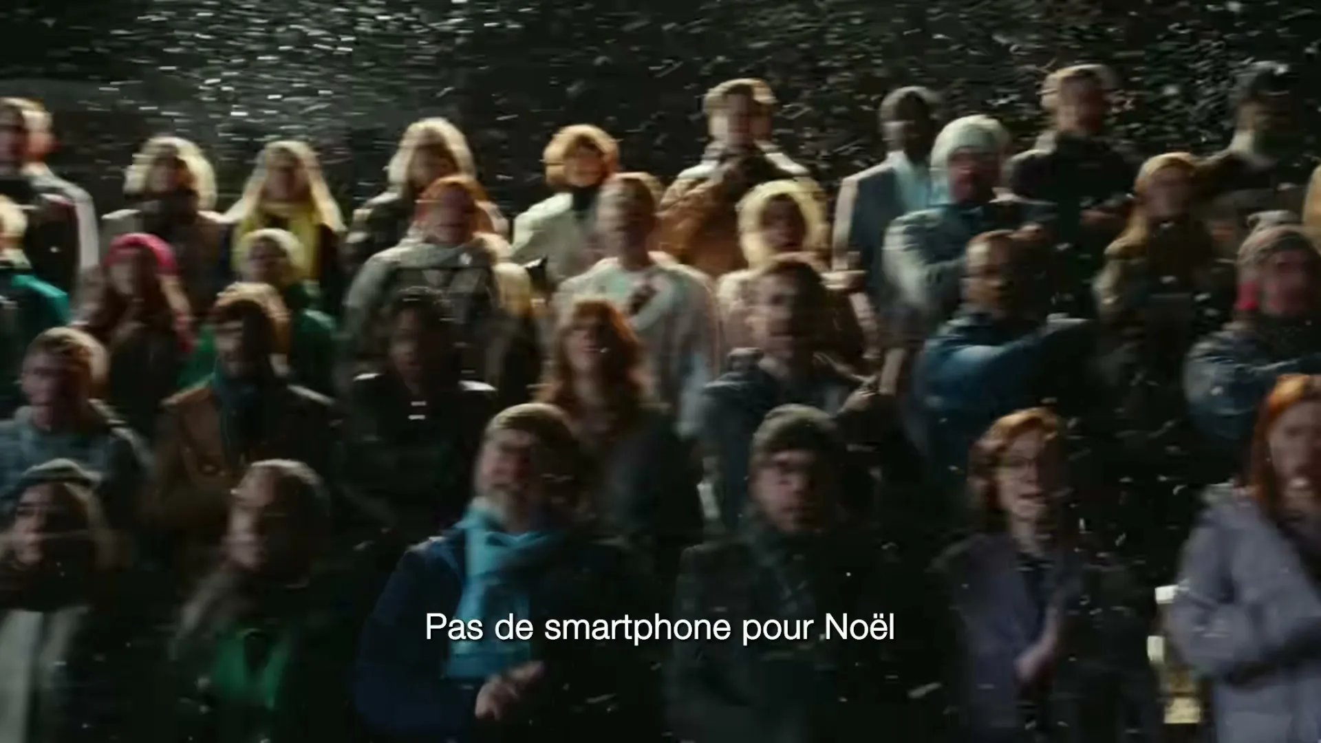 A diverse crowd of people of all ages, dressed in winter clothes, stand together in a dark outdoor setting with snow falling, looking up and singing.