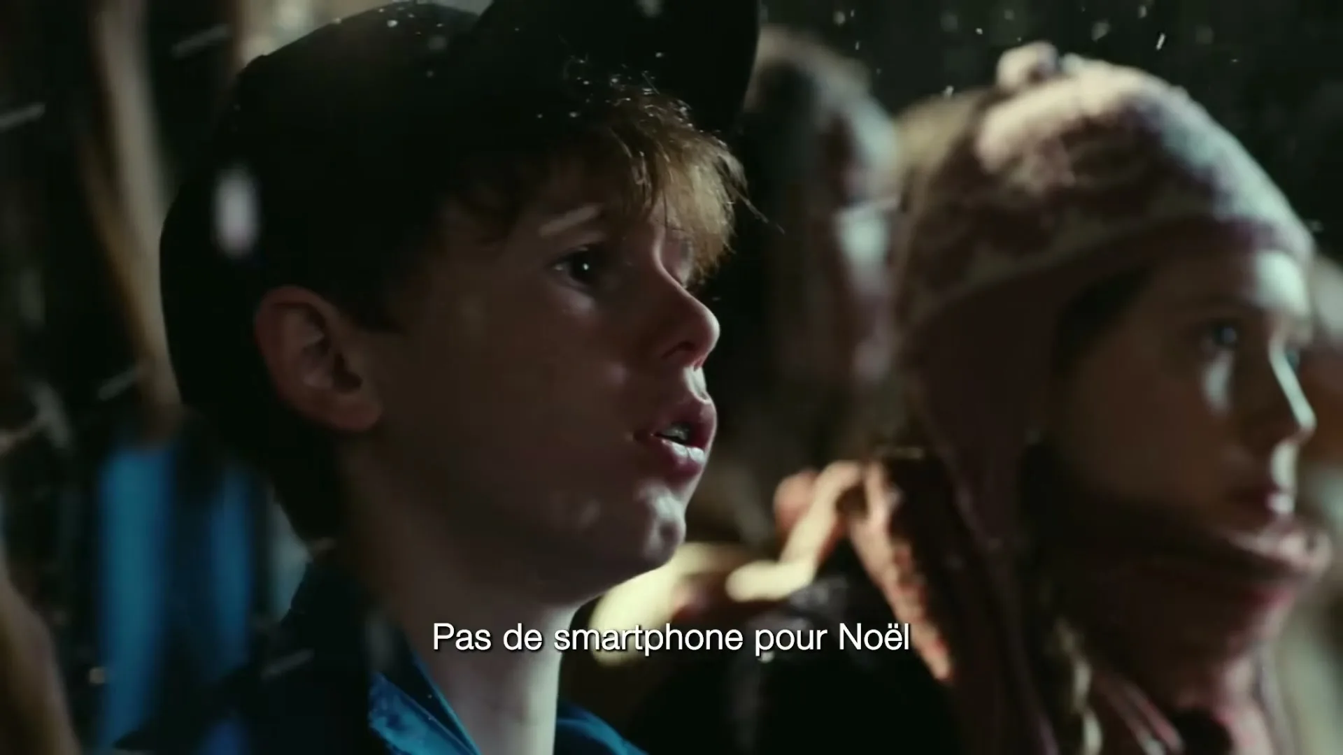A young boy with a black cap and blue jacket looks up in wonder as snow falls around him, with another child partially visible beside him.