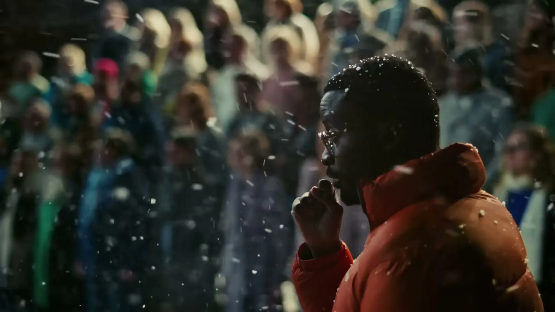 A man in an orange puffer jacket stands in the foreground, looking at a crowd of diverse people in winter clothing, as snow falls around them at night.