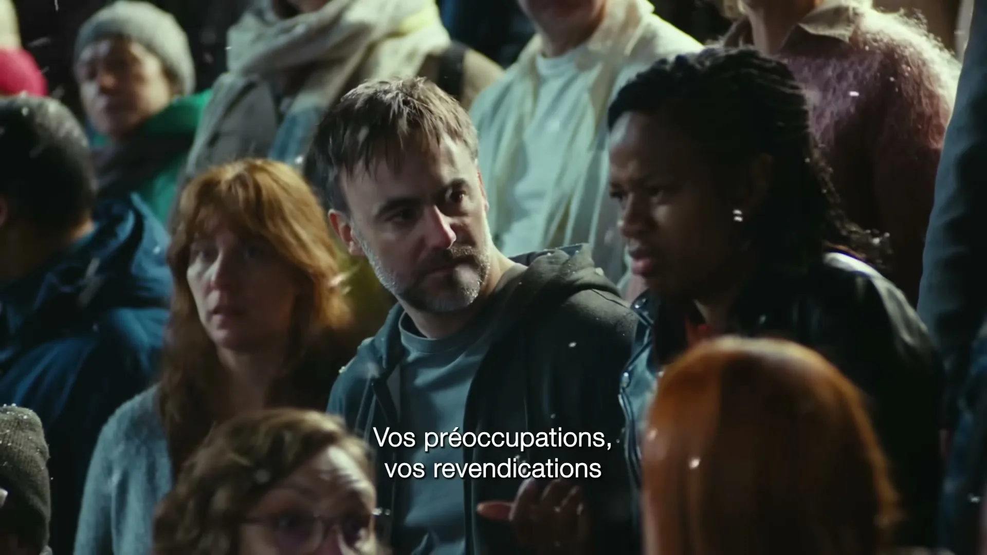 A diverse crowd of people, including a man with a beard and a woman with braids, look on with concerned expressions as snow falls around them in an outdoor setting.
