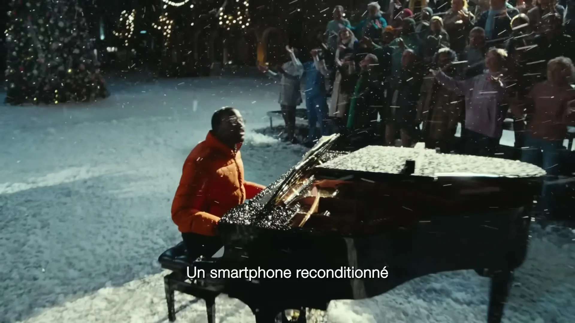 A Black man in an orange puffer jacket plays a grand piano in the snow, surrounded by a cheering crowd at night.
