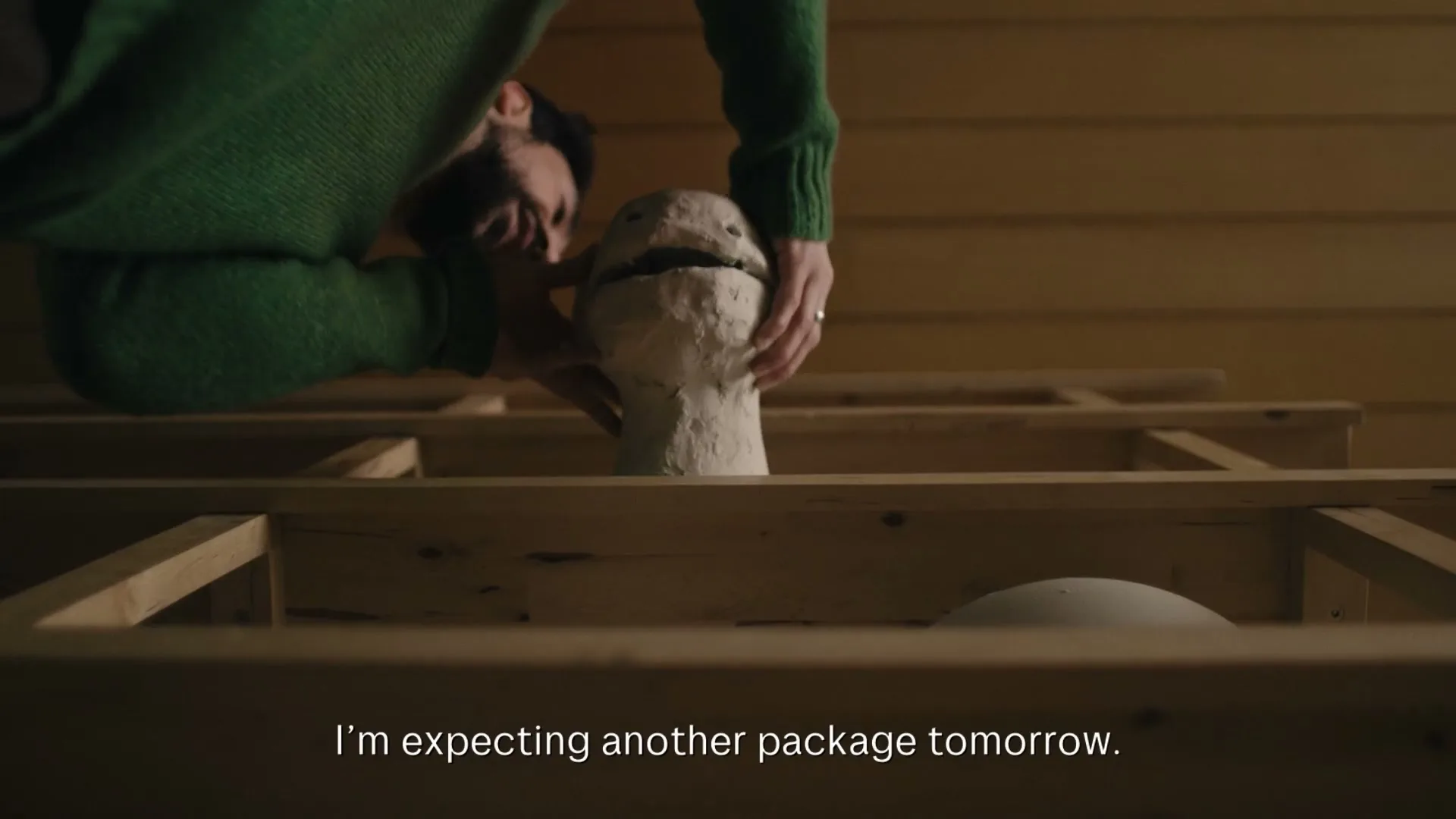 A man in a green sweater is seen from a low angle, working on a clay sculpture of a creature's head, which is emerging from wooden floorboards.