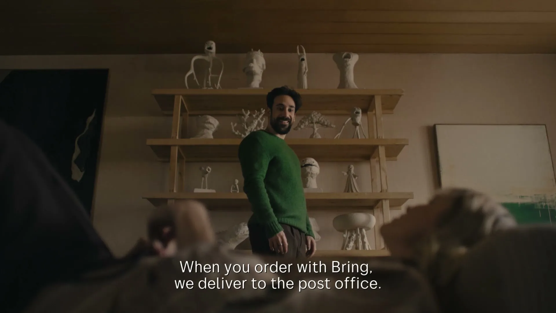 A man in a green sweater stands in a living room with shelves of sculptures, while a woman lies on a couch in the foreground.