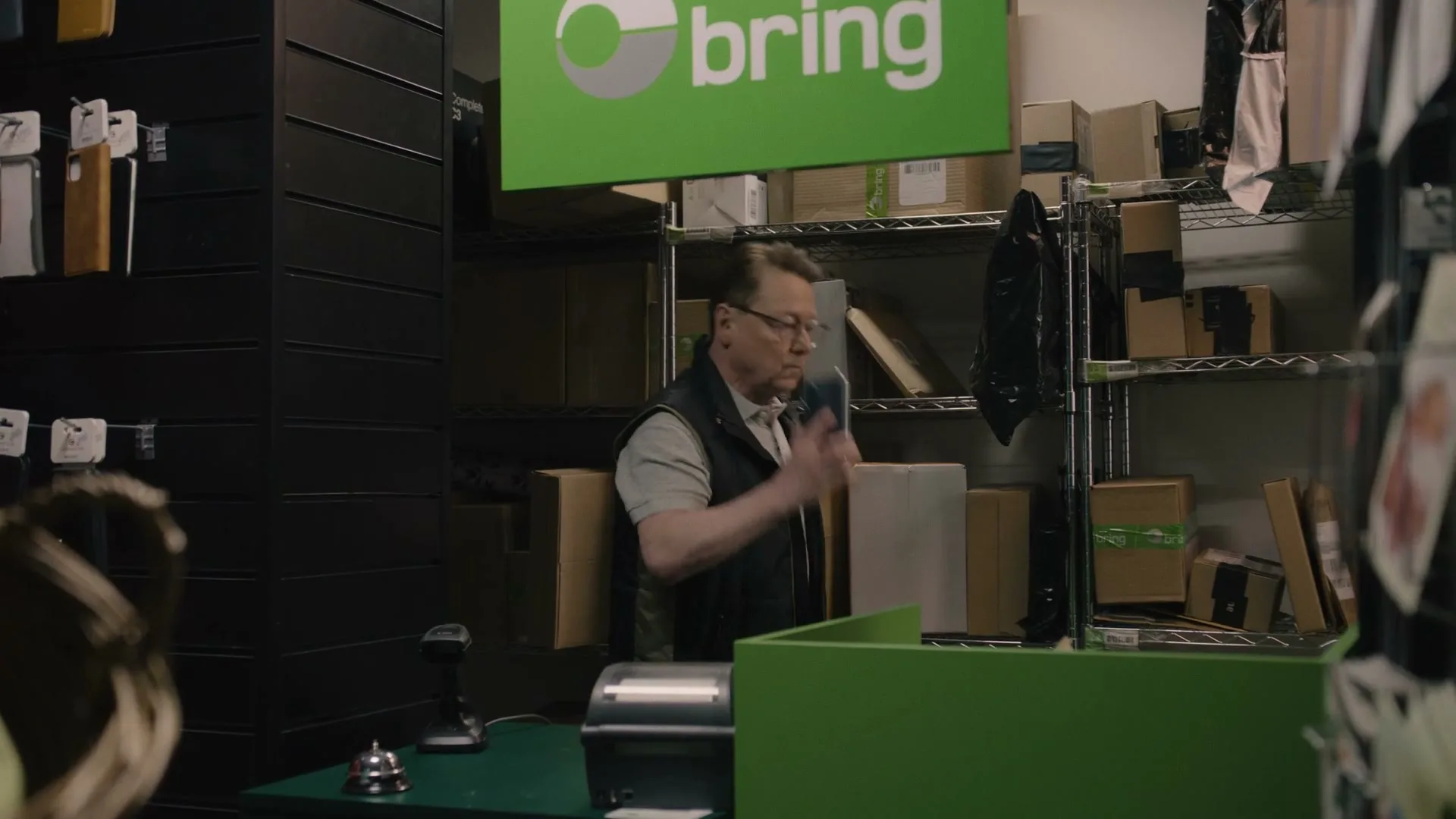 A man in glasses and a vest scans a package at a post office counter, then answers a phone call, looking up in confusion.