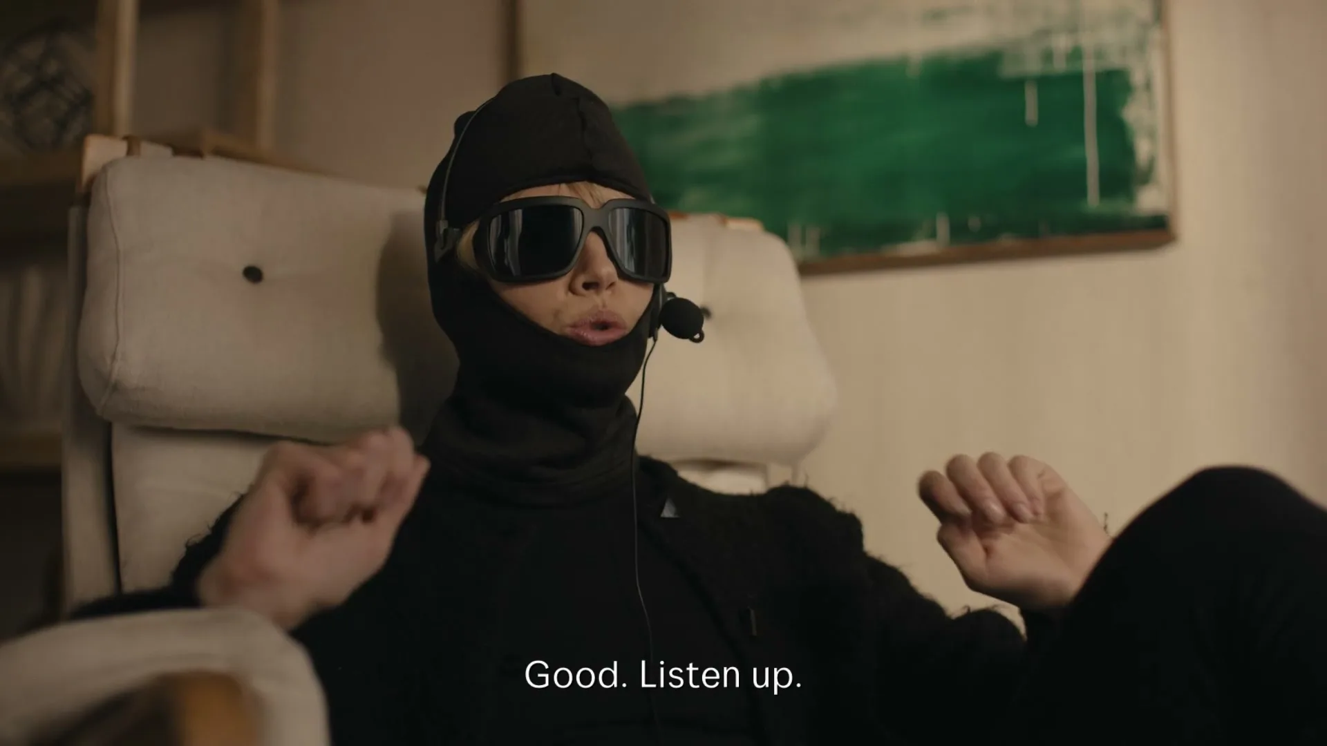 A person in a black balaclava, sunglasses, and headset sits in a chair, talking on the phone in a dimly lit room.