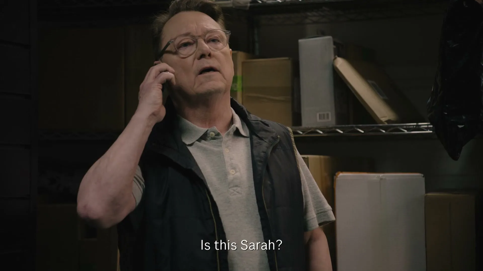 A man in his 60s, wearing glasses and a vest, holds a phone to his ear, looking up in a storage room.