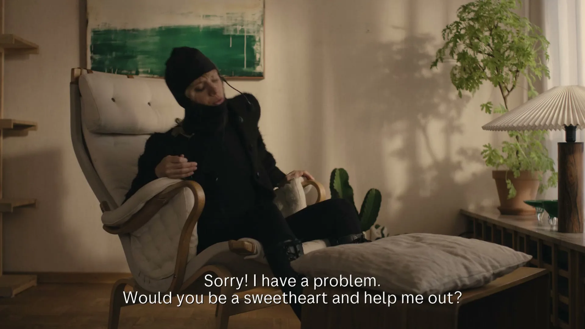 A person in a black balaclava and sunglasses reclines in a chair, talking on a headset, in a cozy living room.