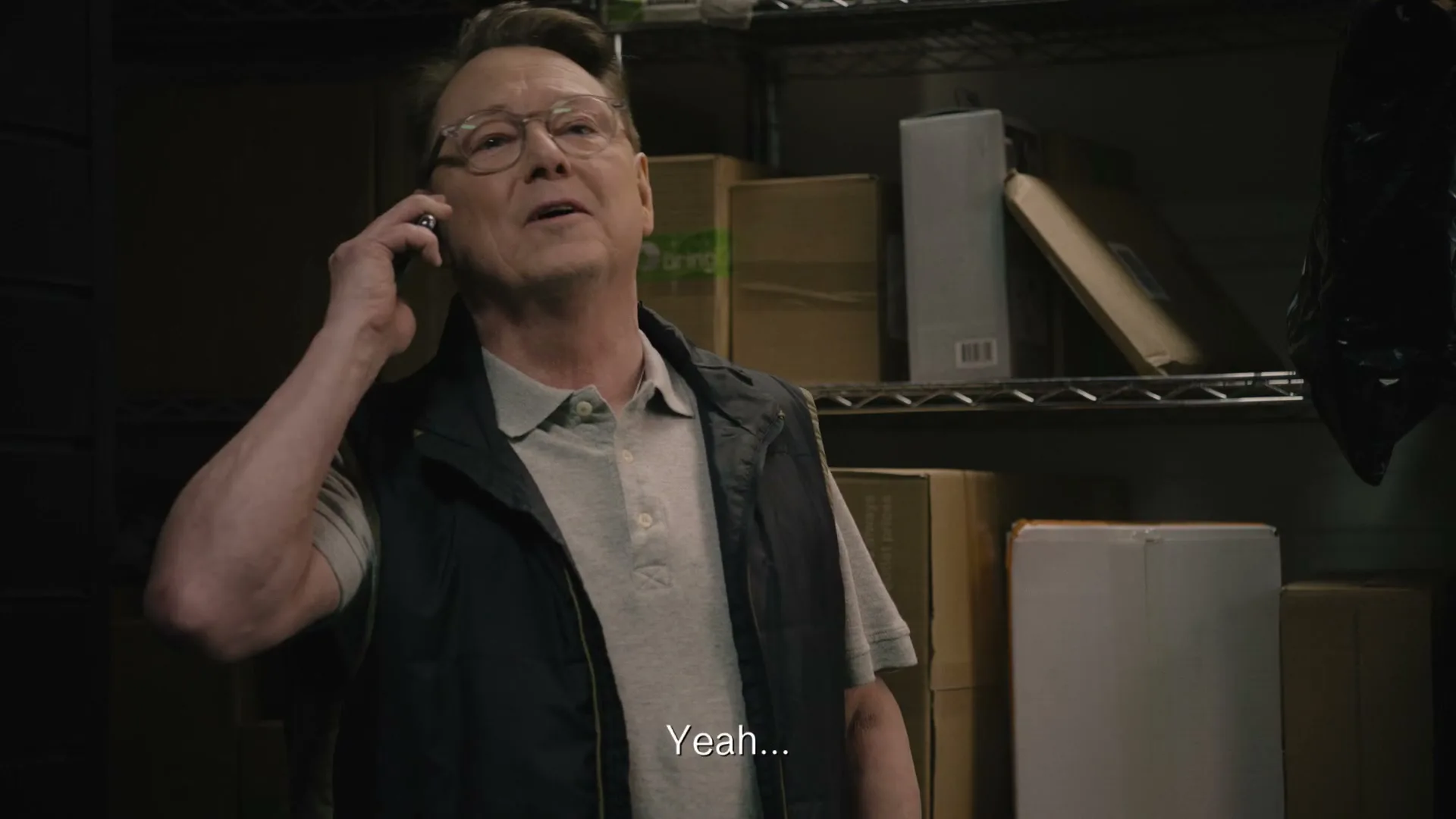 A middle-aged man in glasses and a vest talks on a phone, looking up in a storage room.