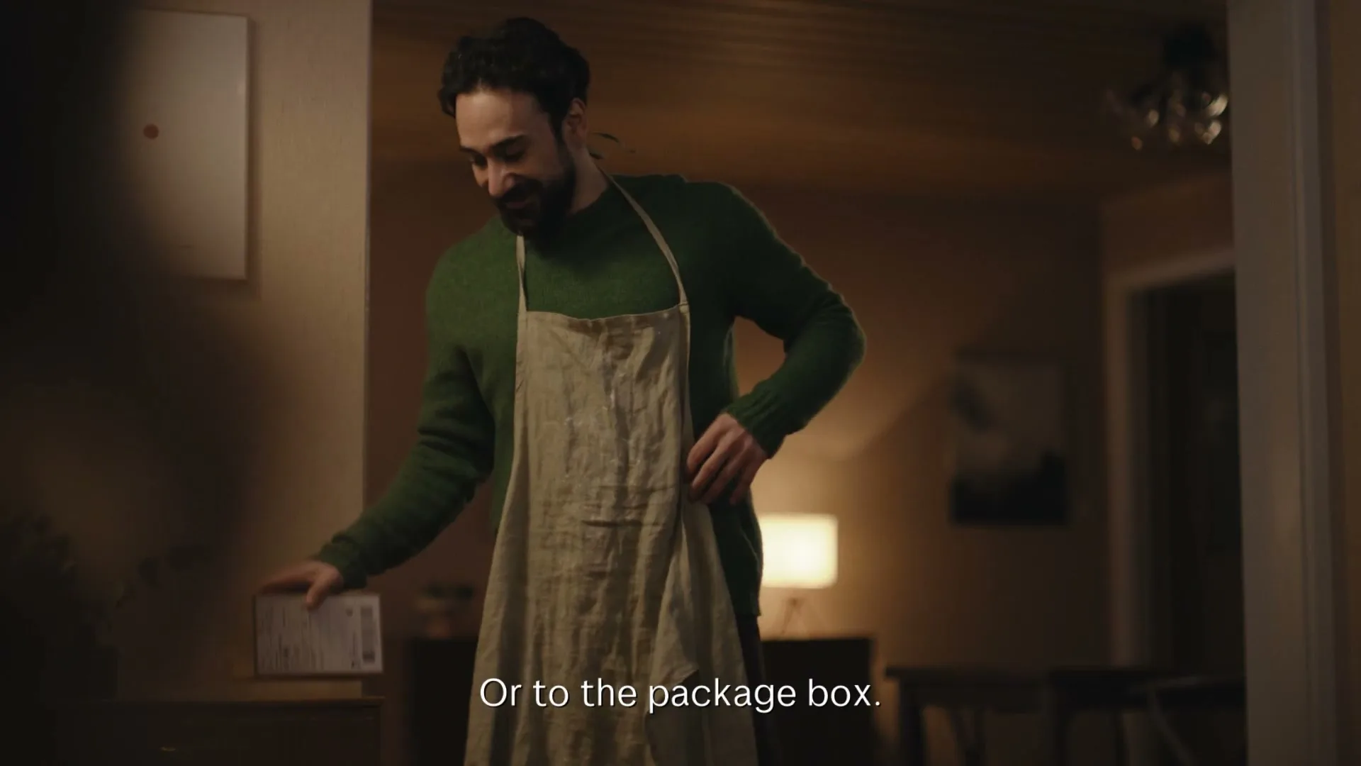 A man in a green sweater and apron holds a package, then puts it down and adjusts his apron in a dimly lit home.