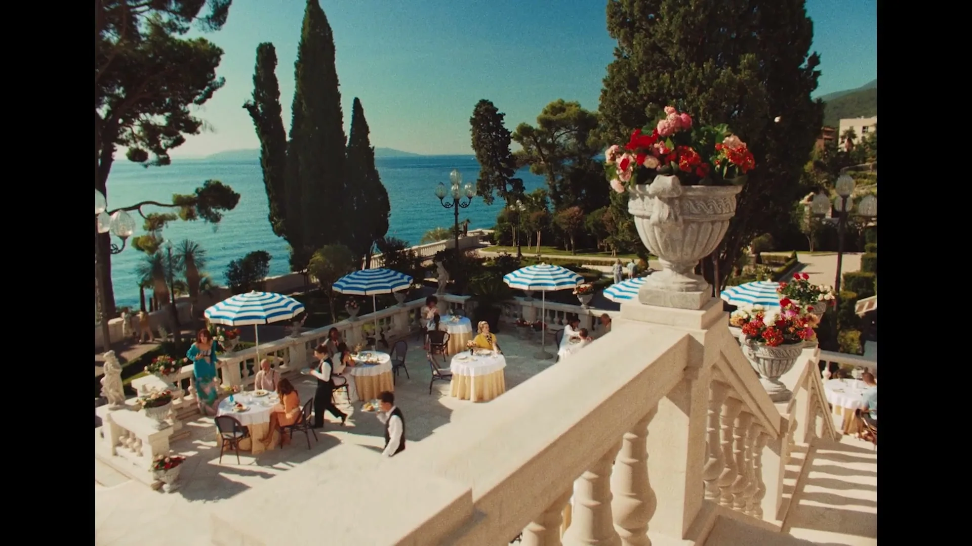 A wide shot of a luxurious outdoor restaurant terrace overlooking the sea, with people dining under striped umbrellas on a sunny day.