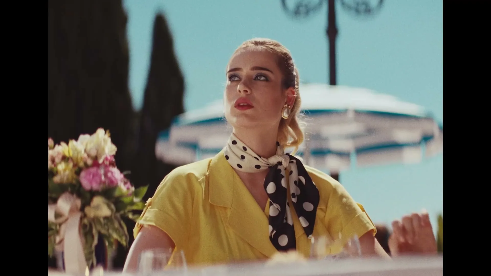 A young woman in a yellow dress and polka dot scarf sits at an outdoor table, looking around with a thoughtful expression on a sunny day.