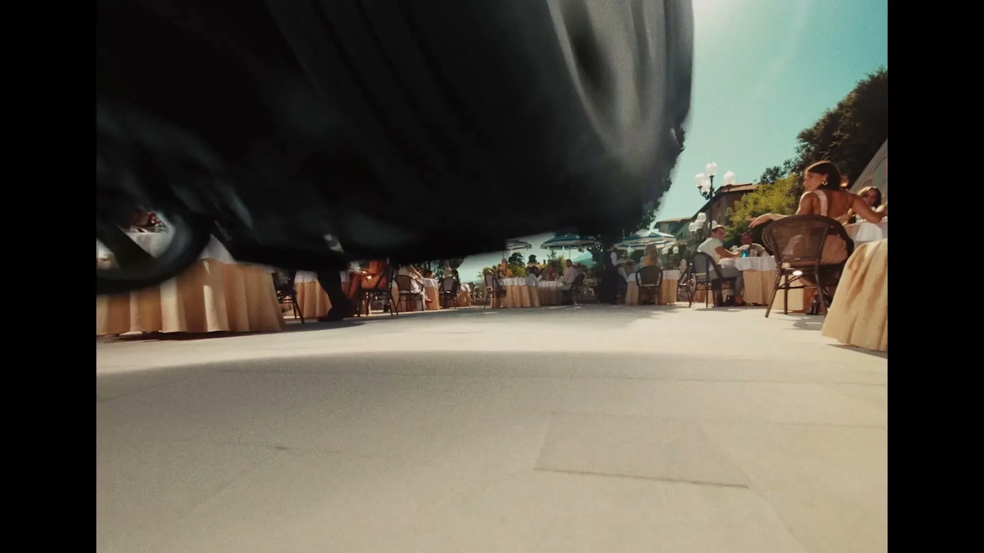 A low-angle shot captures a black car speeding past an outdoor restaurant patio, where people are dining under a bright sky.