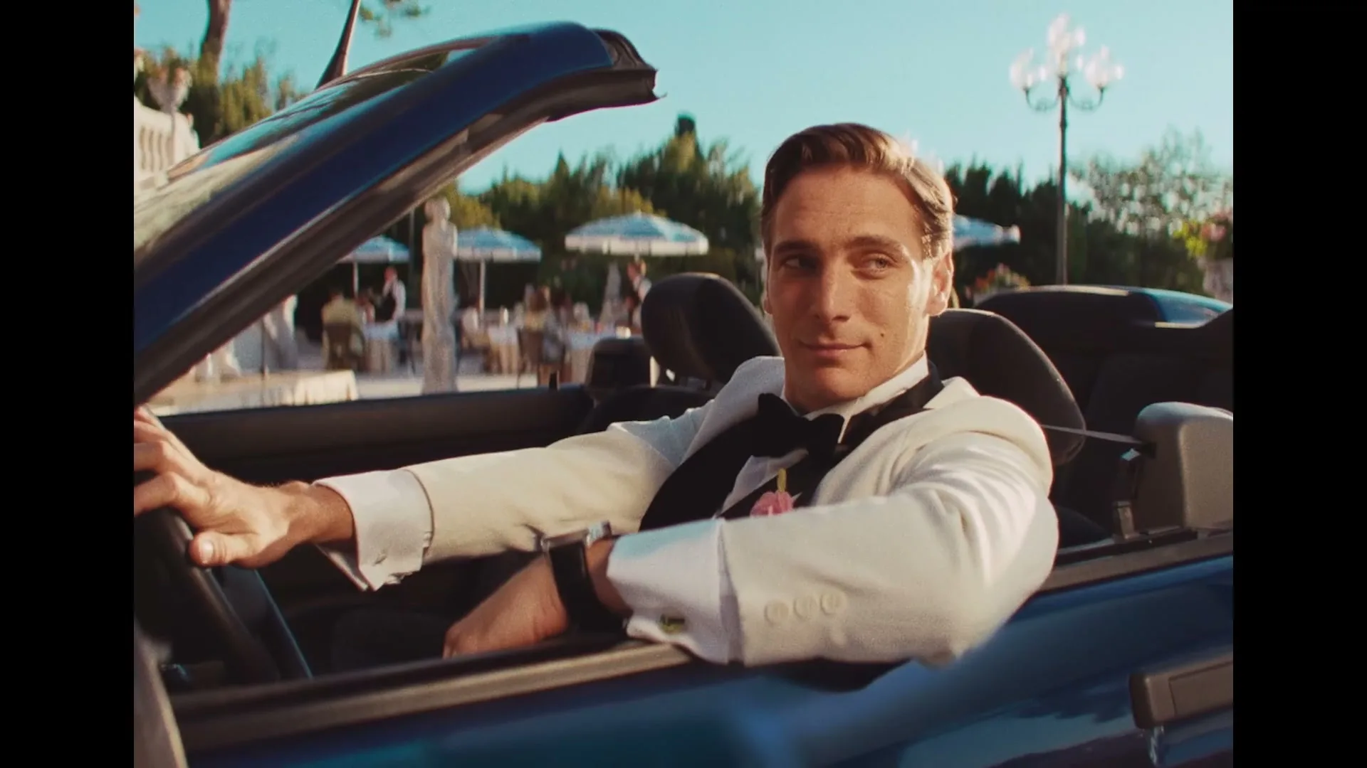 A handsome man in a white tuxedo and black bow tie sits in a blue convertible, looking confidently at the camera in a luxurious outdoor setting.