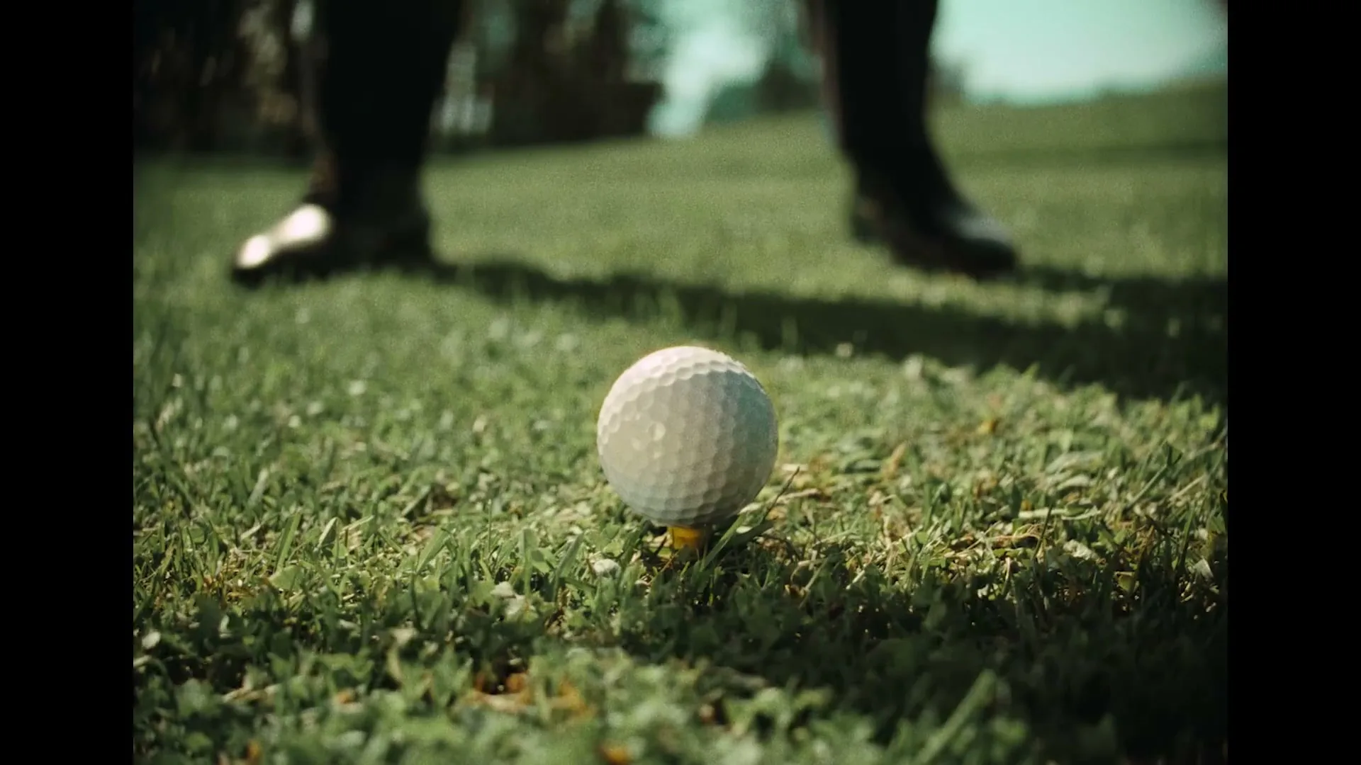 A golf club hits a golf ball off a tee on a grassy golf course, with a person's legs visible in the background.