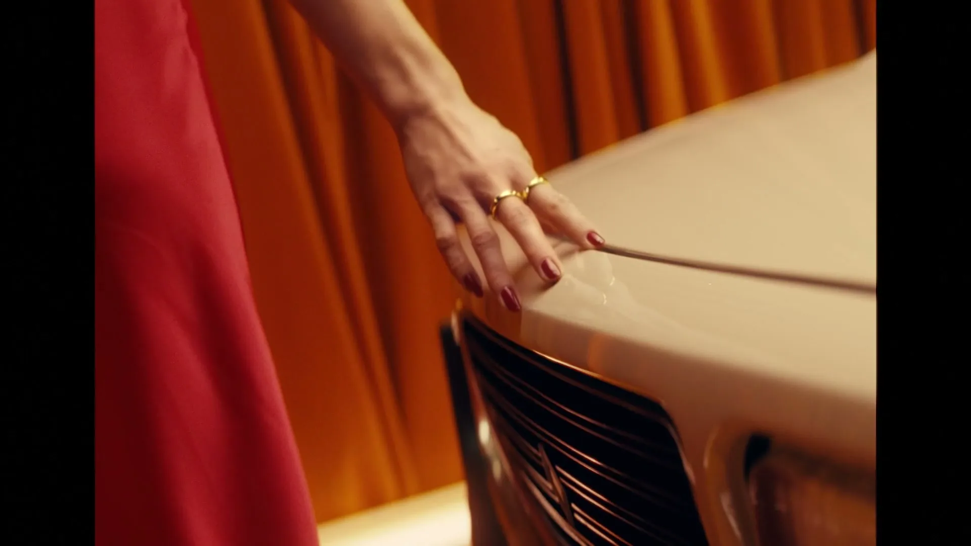 A woman's hand with red nail polish and gold rings gently caresses the hood of a white vintage car, with an orange curtain in the background.