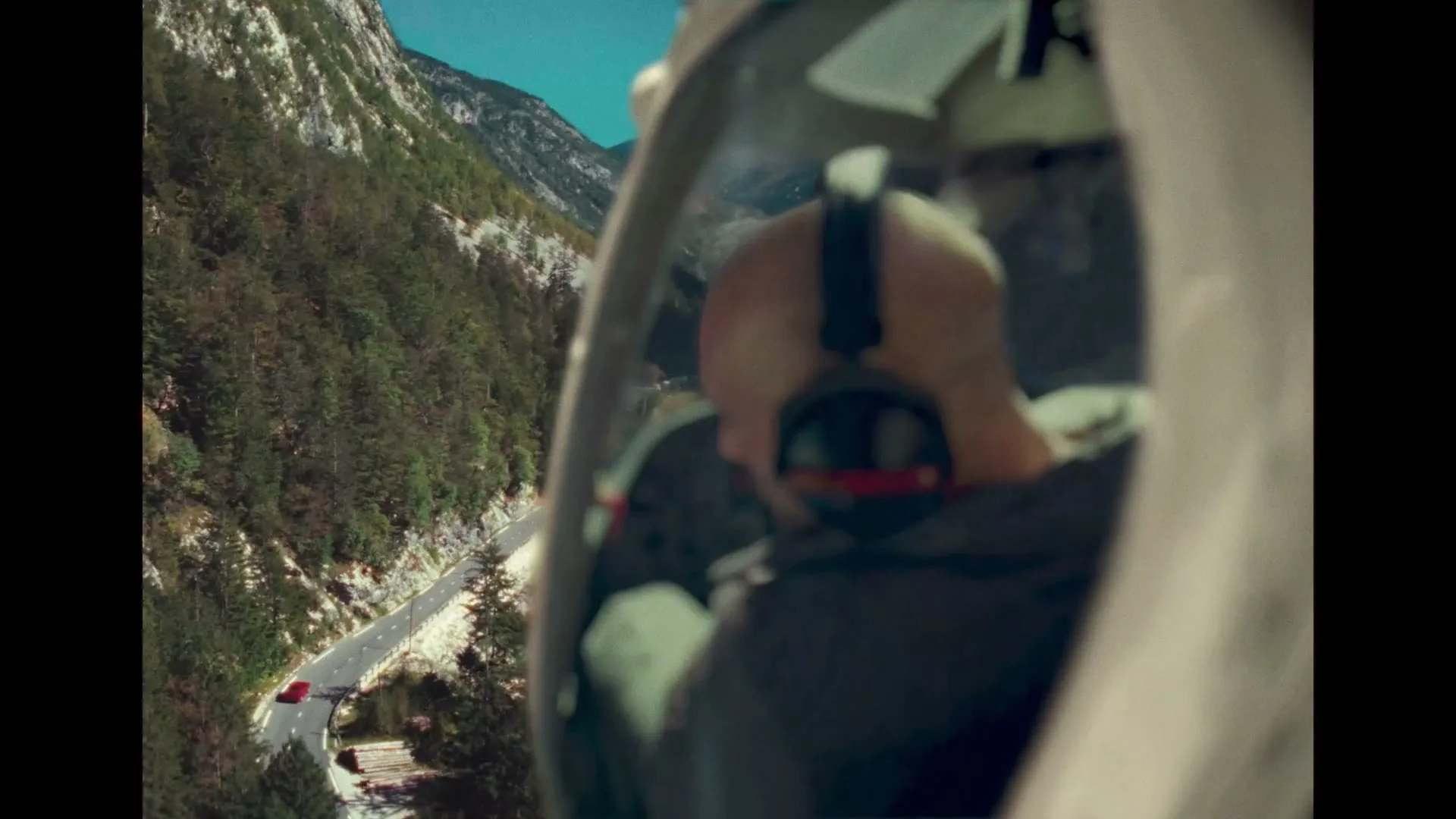 A bald male helicopter pilot in a headset looks down at a red car driving on a winding road through a snowy, forested mountain valley.