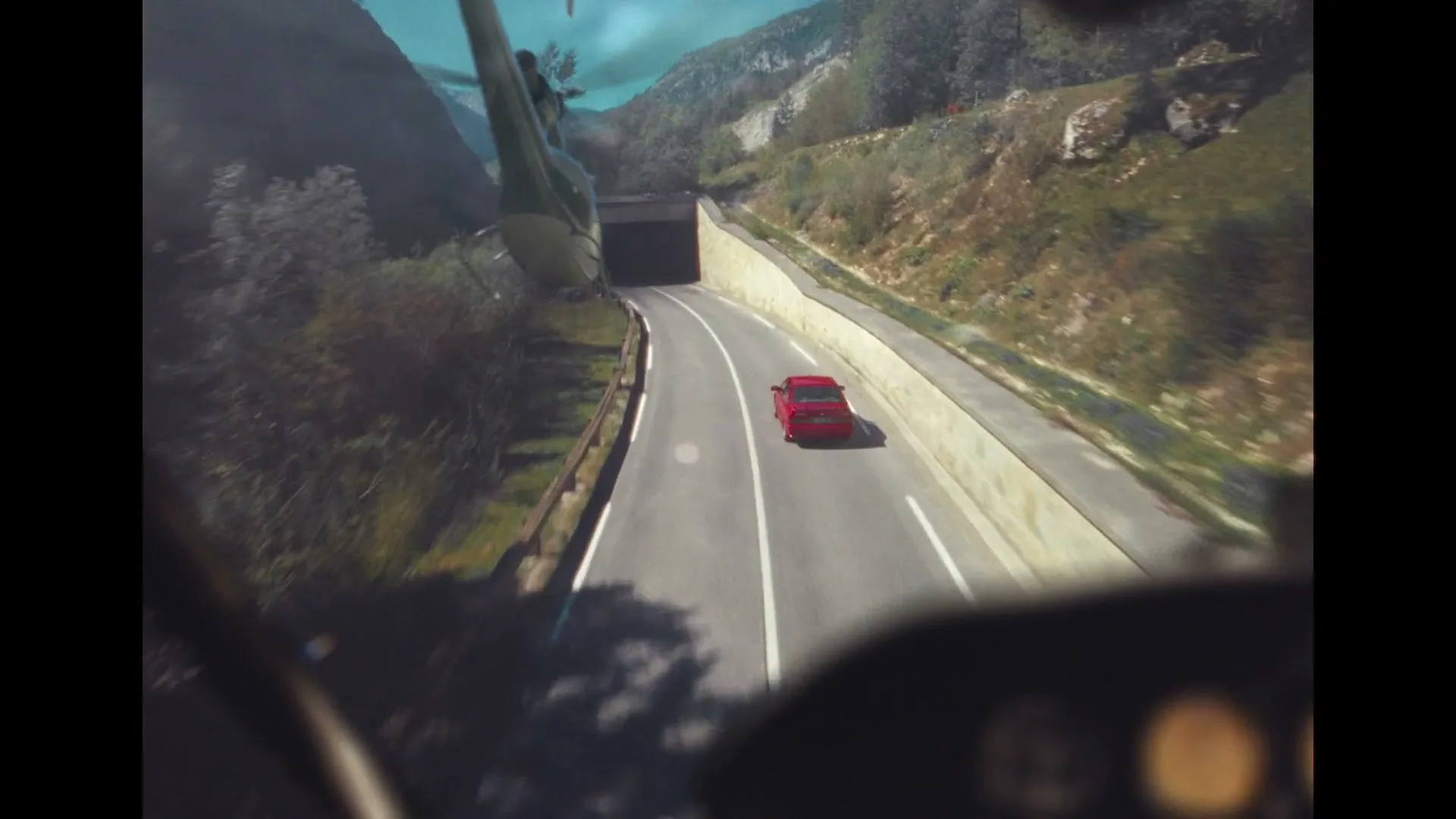 A red car drives on a winding road towards a tunnel, filmed from a helicopter's perspective in a mountainous landscape.