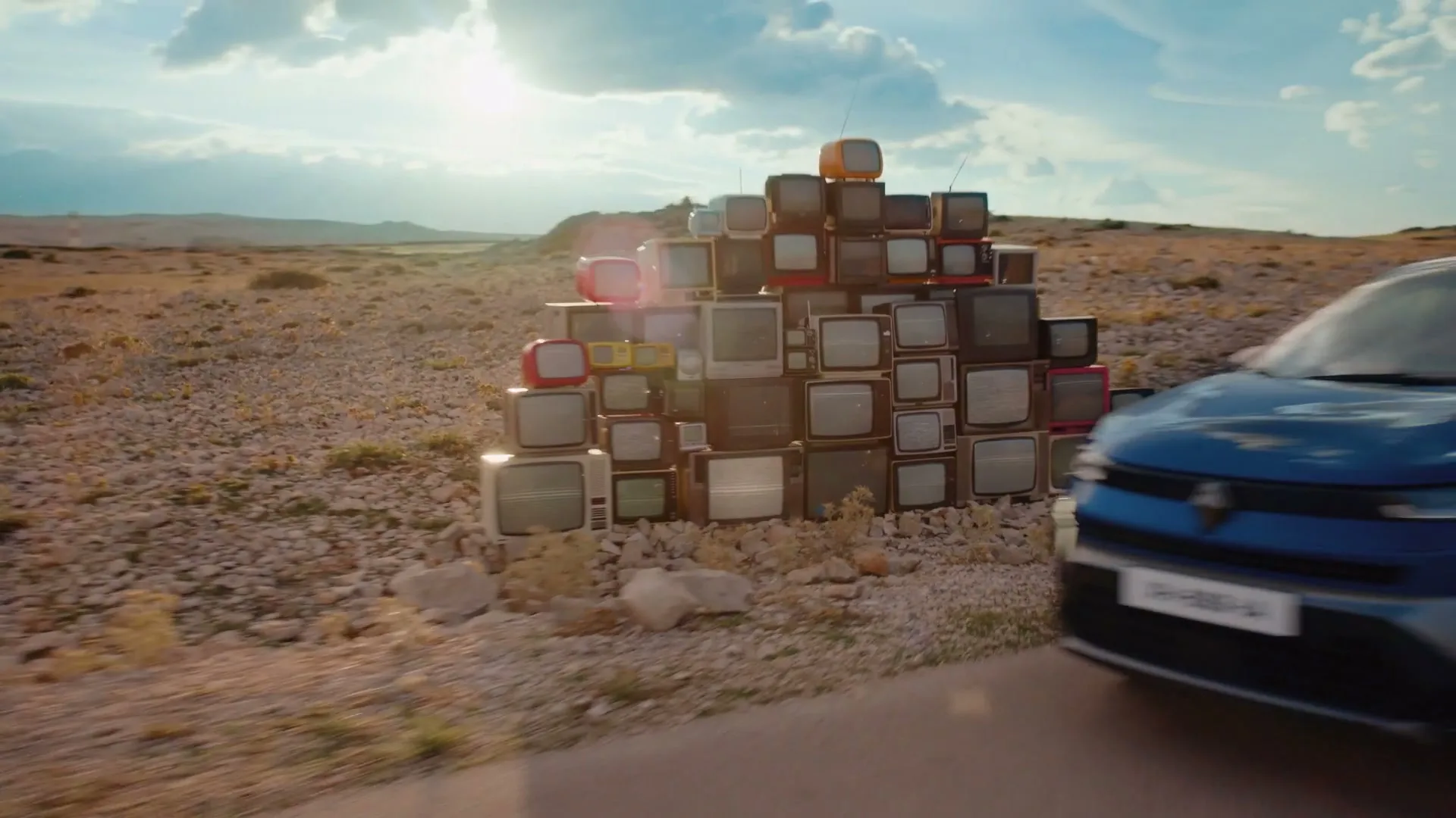 A blue Renault car drives past a large pile of old televisions in a vast, arid desert landscape under a bright, cloudy sky.