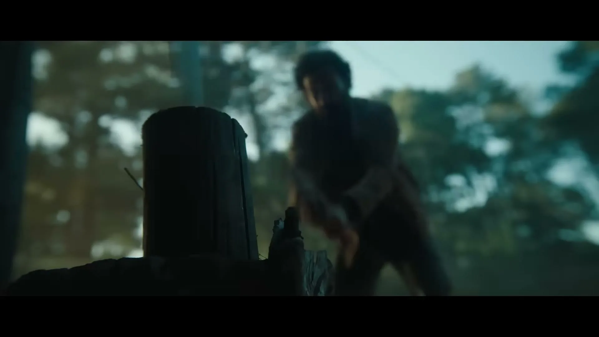 A man in a brown shirt is chopping wood with an axe in a forest clearing during the day.