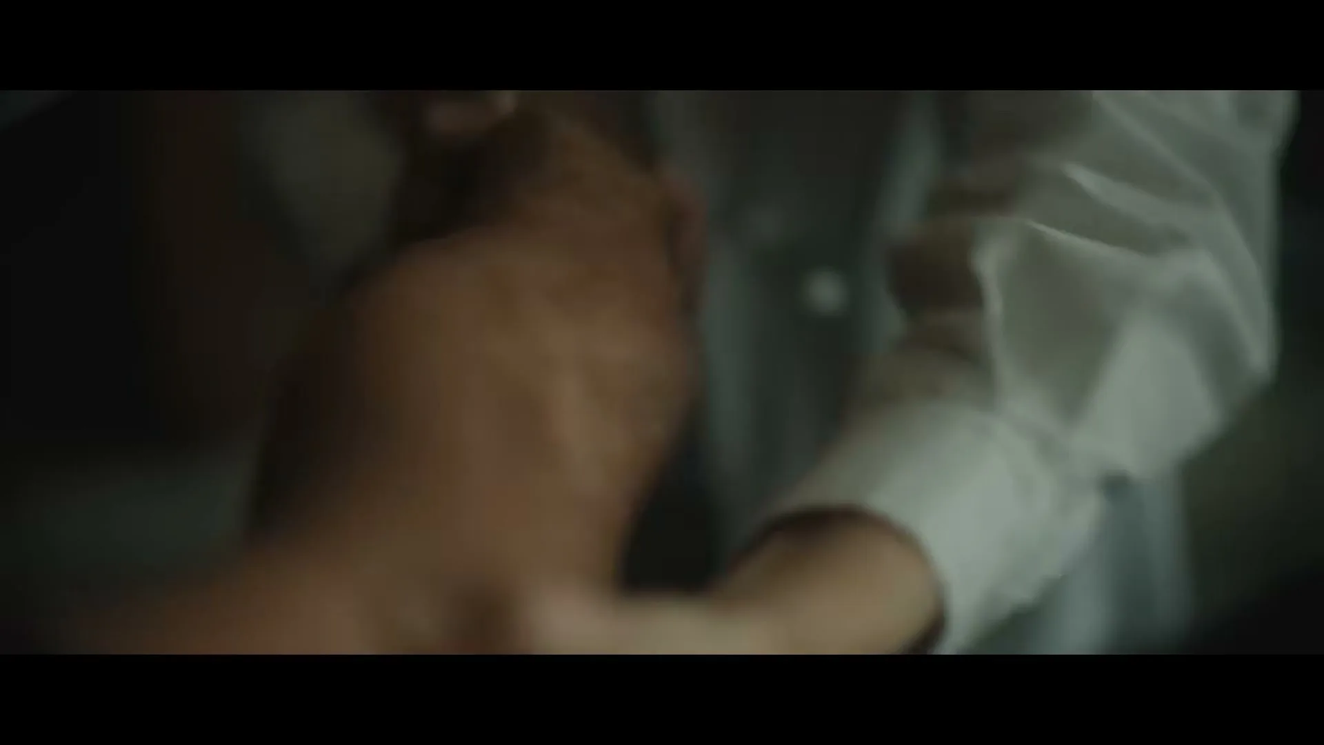 A doctor in a white coat examines the arm of a patient, holding their wrist gently in a dimly lit interior.