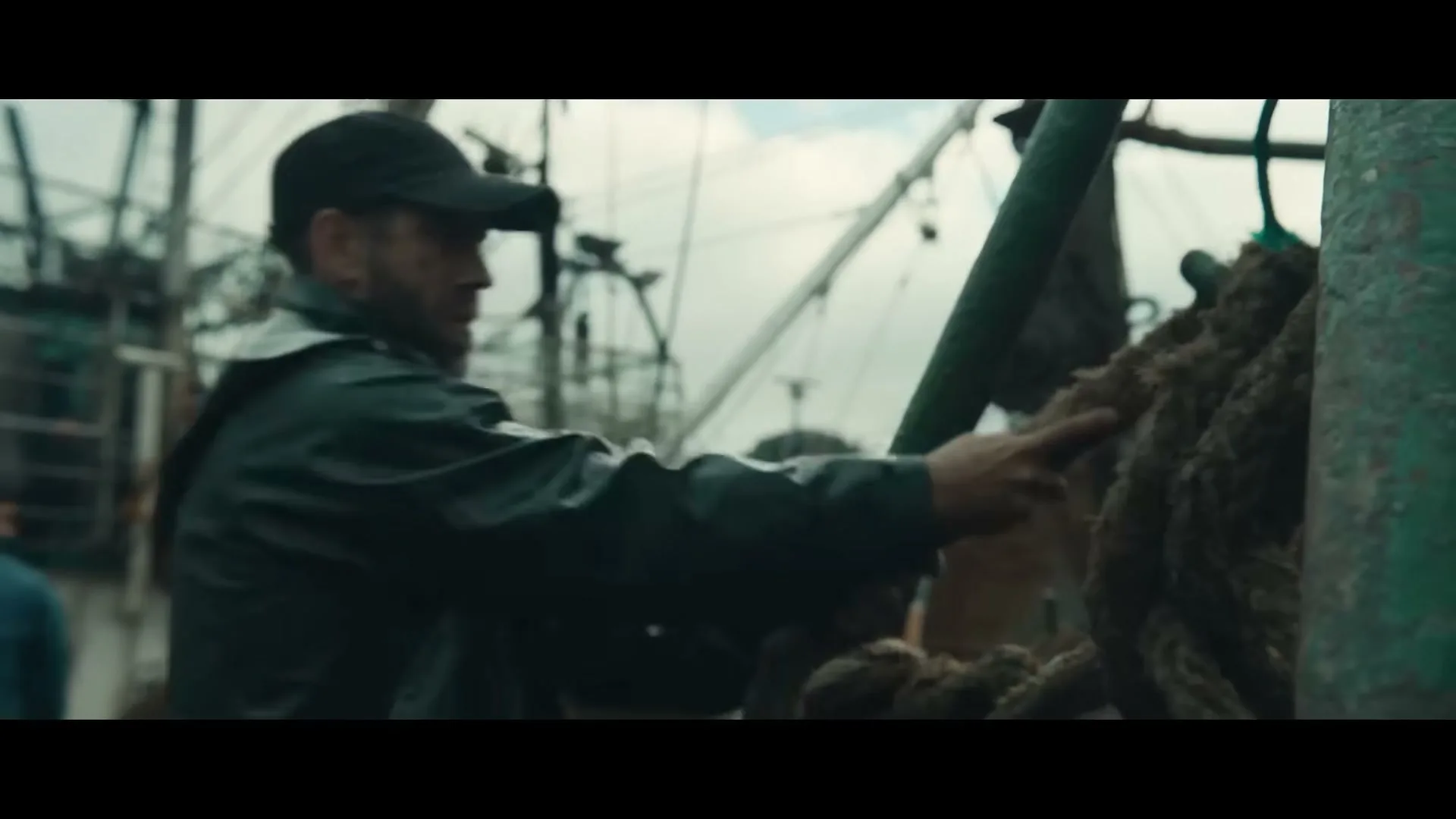 A male fisherman in a dark rain jacket and cap works with thick ropes on a fishing boat under an overcast sky.