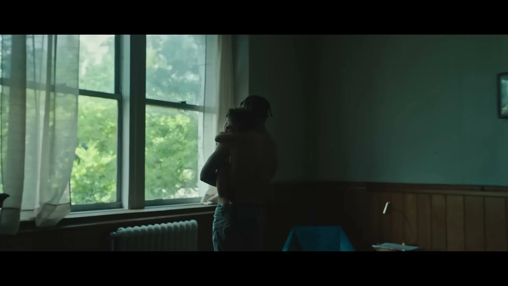 A shirtless man holds a child in his arms, looking out a large window with sheer curtains in a dimly lit room.