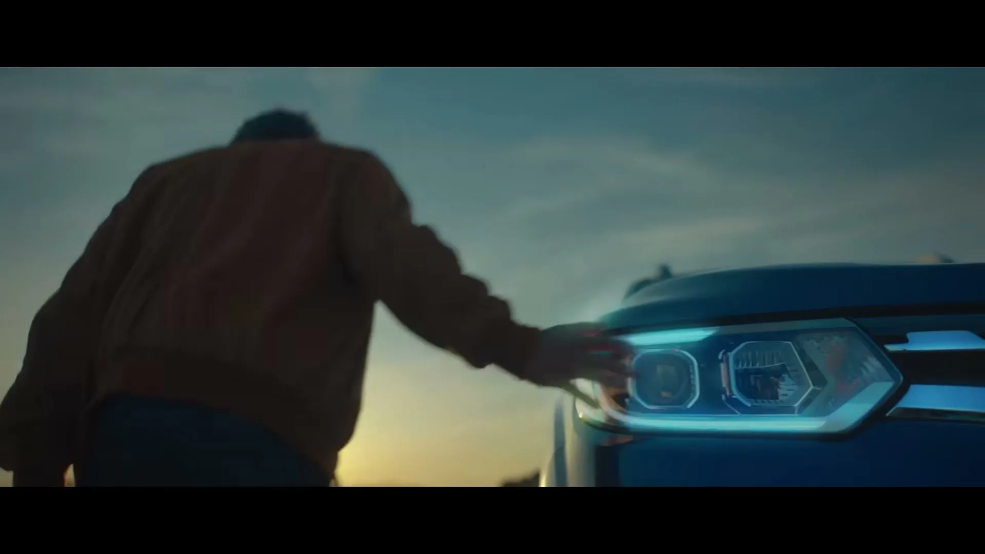 A person in a brown jacket walks away from a blue car, touching its glowing headlight at dusk.
