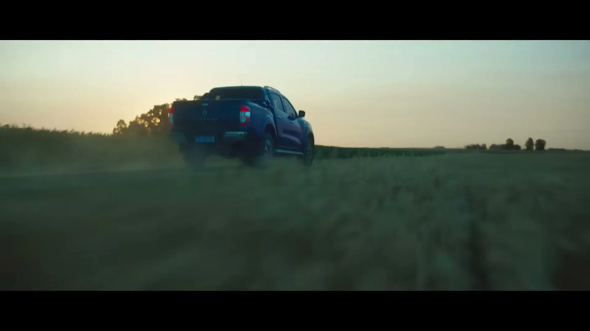 A dark blue pickup truck drives away from the camera through a field at sunset, kicking up dust.