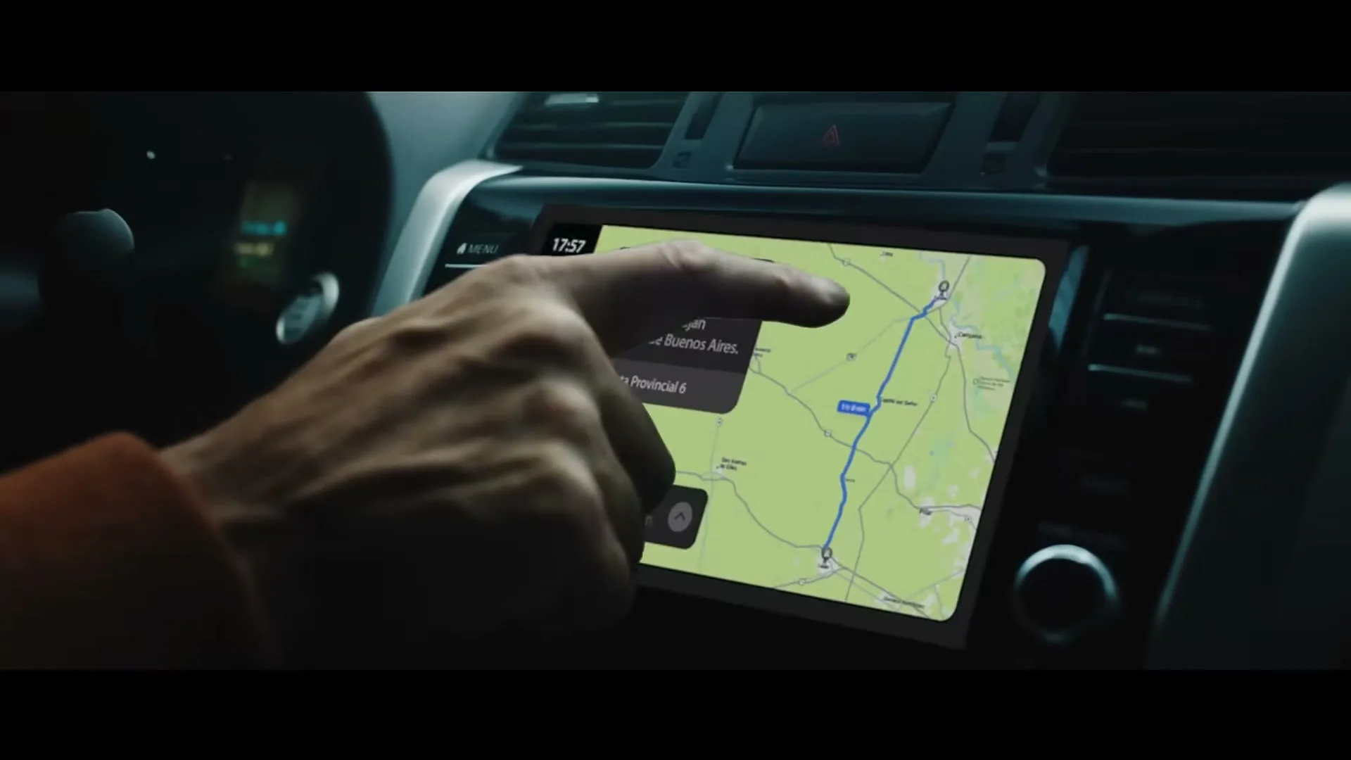 A man's hand taps on a car's infotainment screen, selecting a map application to display a route.