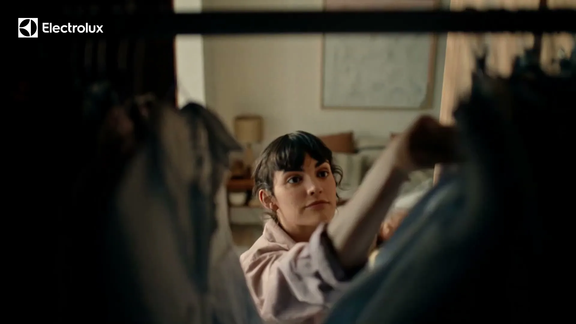 A young woman with dark hair and bangs, wearing a light pink shirt, looks through clothes on a rack in a bedroom.