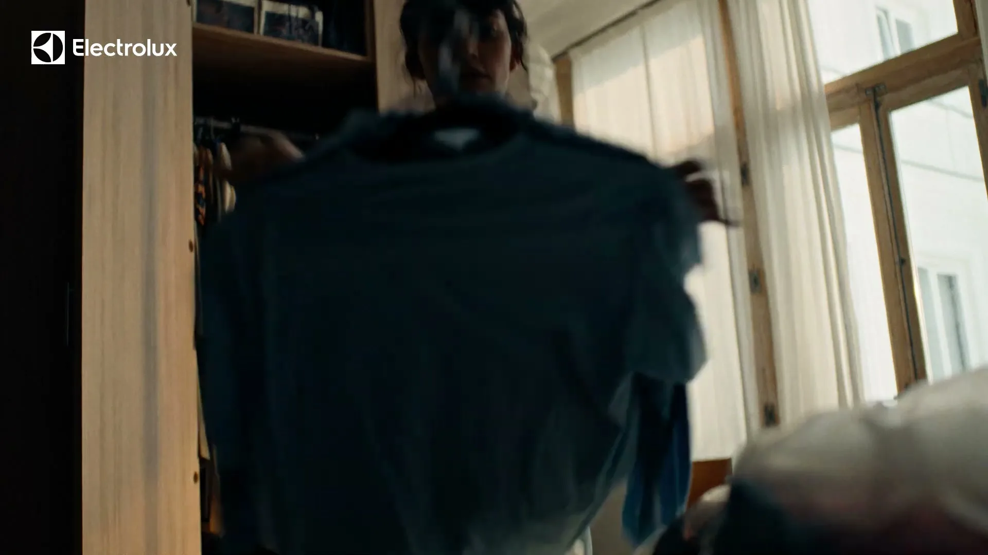 A young woman with short dark hair holds up a light blue t-shirt in front of a closet and a window, then drops it.