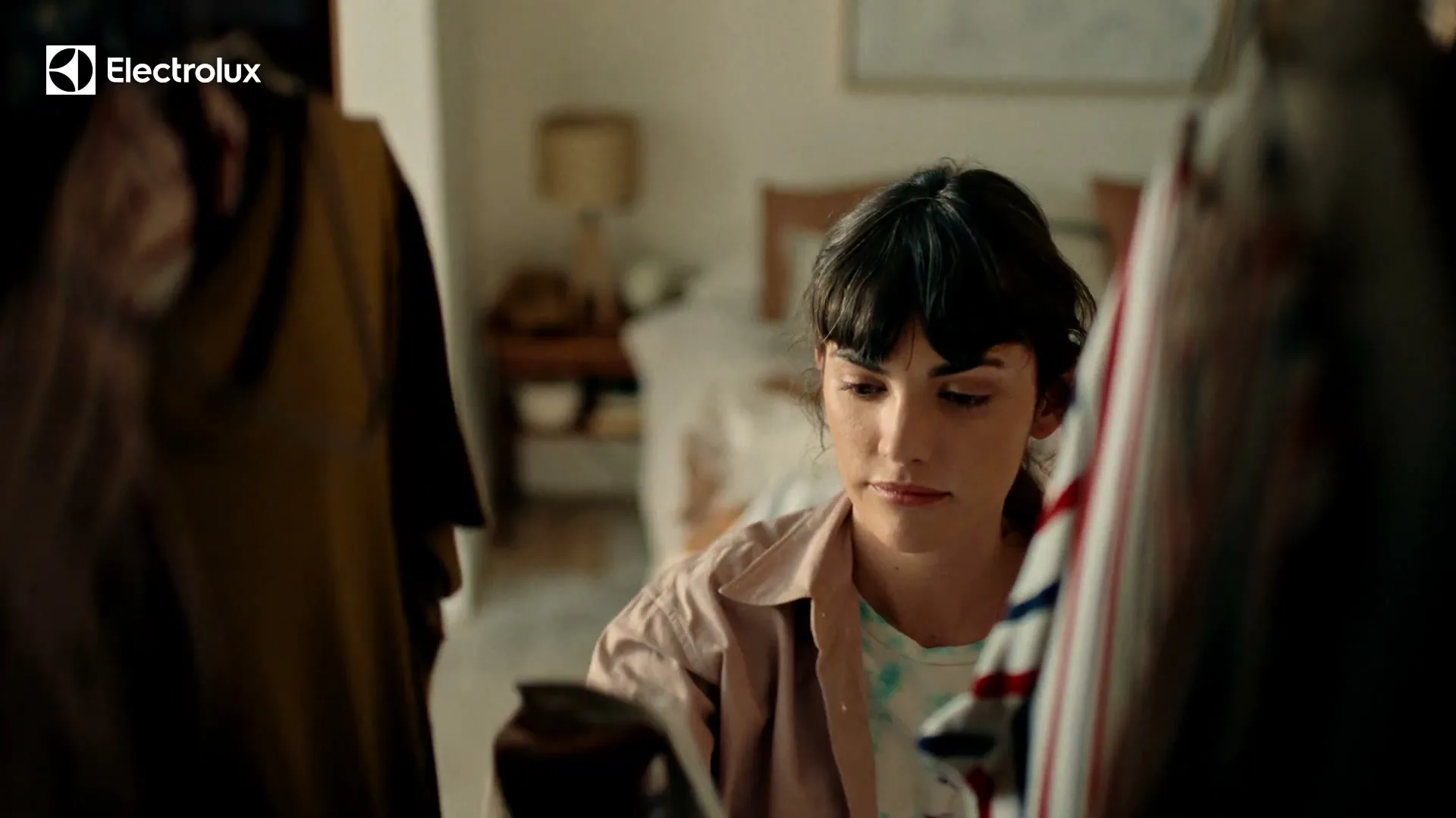 A young woman with dark hair and a pink shirt looks at clothes in a messy bedroom, appearing thoughtful.