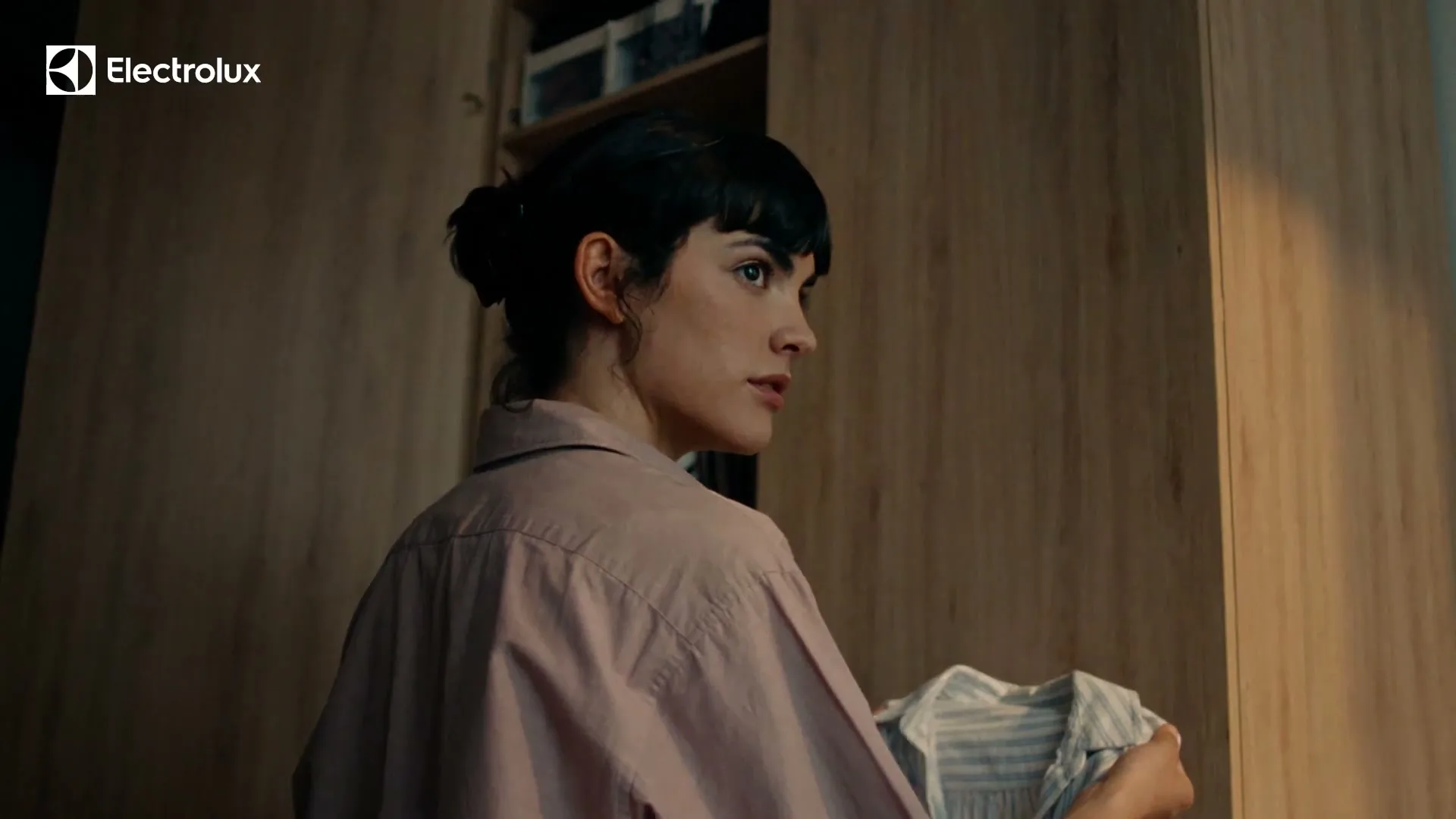 A young woman with dark hair in a bun, wearing a pink shirt, holds a striped shirt while looking into a wooden closet.