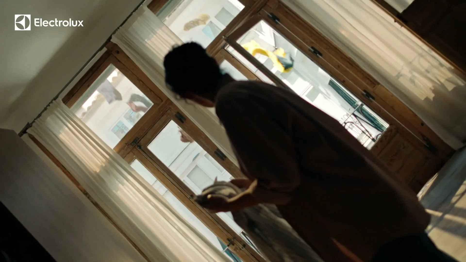 A woman in her 30s, seen from behind, stands in a sunlit room, holding a small item, looking out through large windows at a building across the street.