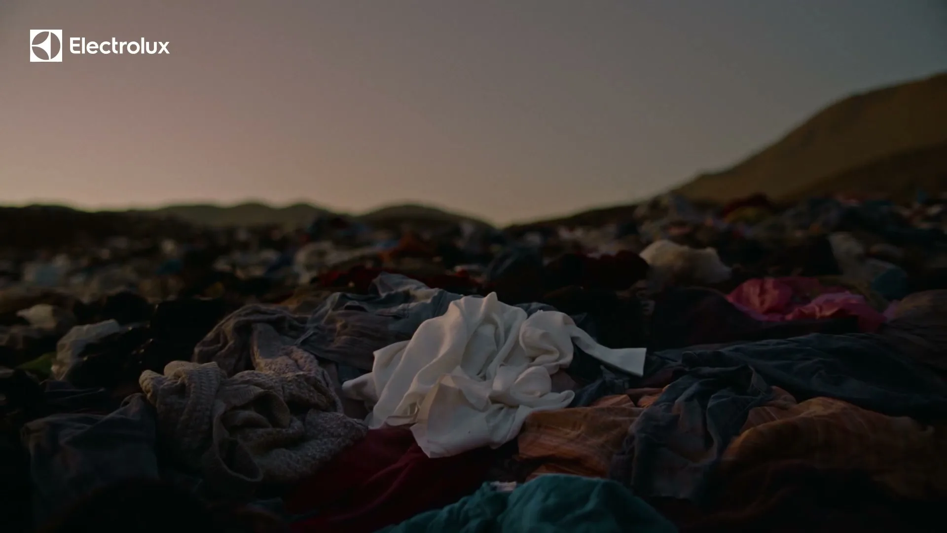 A wide shot shows a vast landfill of discarded clothes under a dusky sky, with a white garment prominently featured in the foreground.