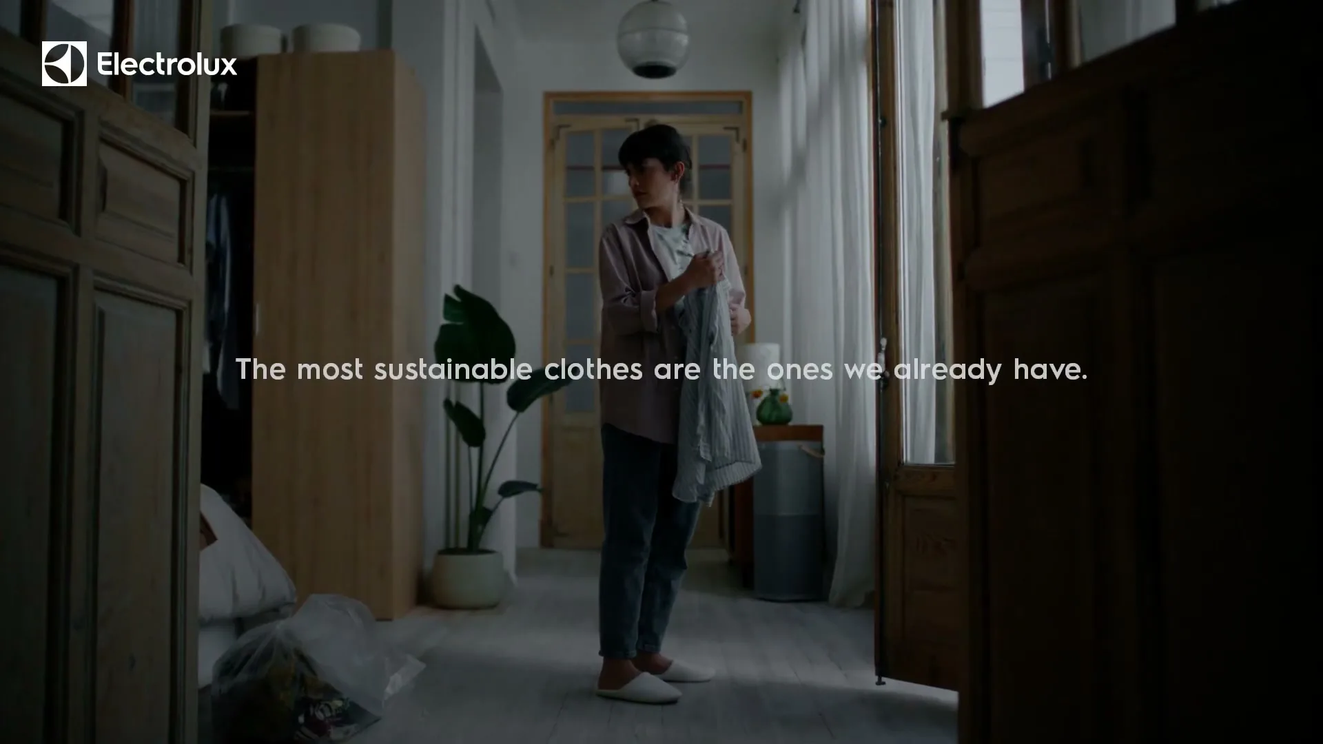 A young woman stands in a hallway, holding a piece of clothing, looking around thoughtfully in a dimly lit home.